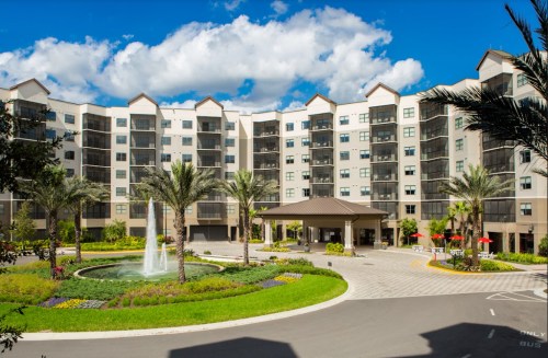 Modern apartment building with a fountain, palm trees, and landscaped gardens under a blue sky with fluffy clouds.