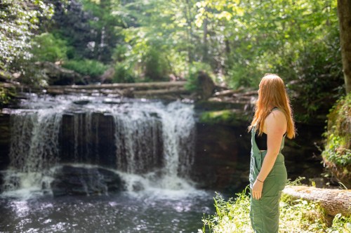 A person with long red hair stands by a serene waterfall surrounded by lush greenery.