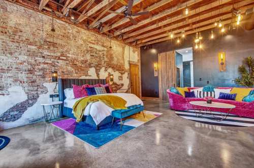 A vibrant bedroom with exposed brick walls, colorful decor, a cozy bed, and a stylish seating area.