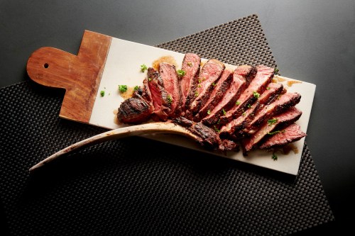 Sliced steak arranged on a wooden cutting board, garnished with herbs, on a dark textured surface.