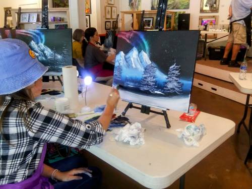 A person paints a snowy mountain scene with northern lights at an art event, surrounded by other attendees.