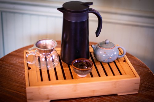 A wooden tray holds a black pitcher, a glass teapot, a small bowl of tea leaves, and a blue teapot.