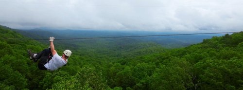 A person zip-lining over a lush green forest with mountains and cloudy skies in the background.