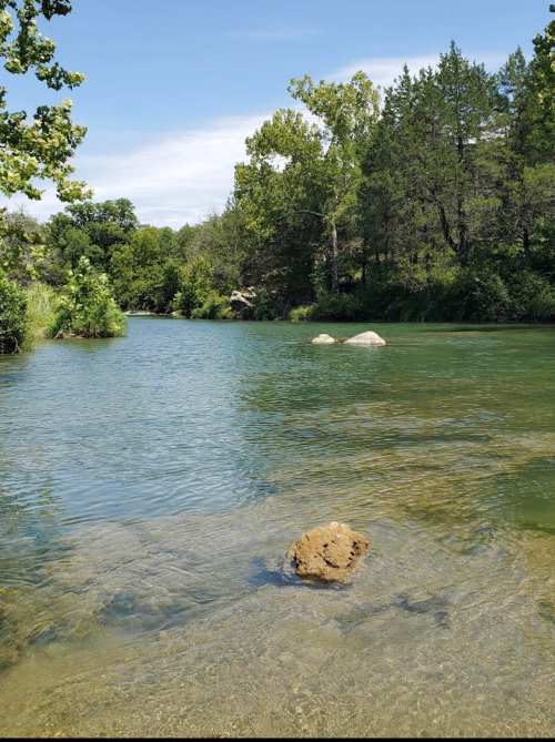 A serene river scene with clear water, rocks, and lush green trees lining the banks under a blue sky.