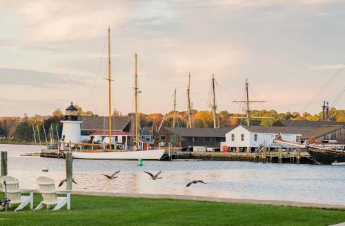 A scenic harbor with a lighthouse, sailboats, and geese flying over calm waters, surrounded by autumn foliage.