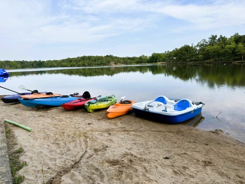 Colorful kayaks and a paddle boat lined up on a sandy shore by a calm lake, surrounded by greenery.