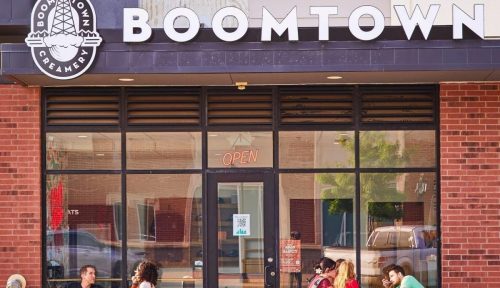 A group of people enjoying drinks at outdoor tables in front of a café named Boomtown, with a welcoming open sign.