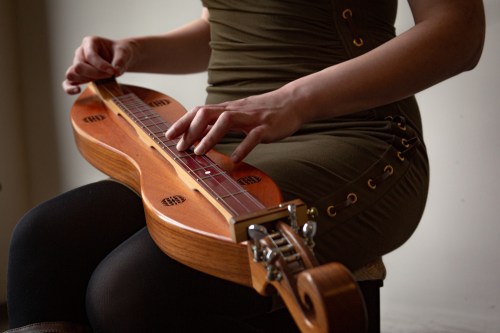 A person plays a wooden dulcimer while seated, wearing a fitted green dress.