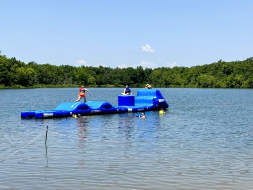 A blue inflatable water play structure in a lake, with people enjoying activities on and around it under a clear sky.