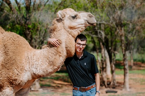A man in a black polo shirt stands next to a camel, gently resting his arm on its neck in a natural setting.