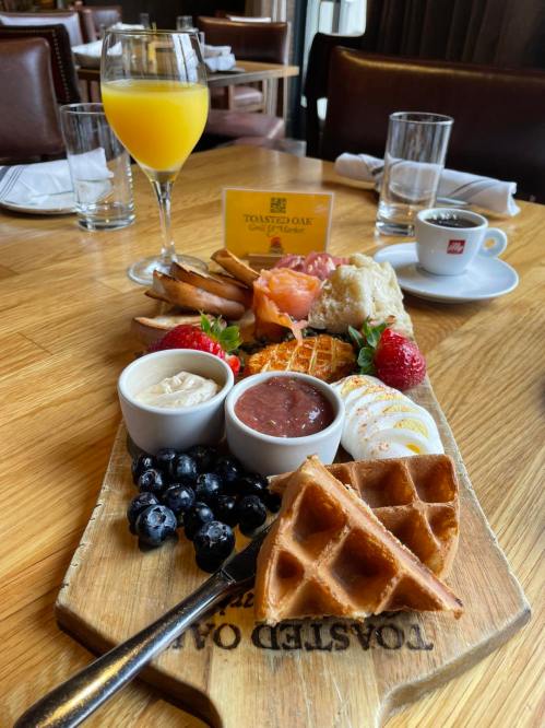 A wooden platter with waffles, fruit, dips, and a mimosa on a table in a restaurant setting.