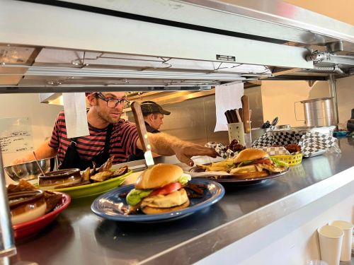 A busy kitchen scene with two chefs preparing burgers and dishes on a counter, showcasing colorful plates of food.