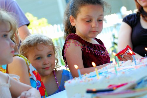 Two young girls watch a birthday cake with candles, one looking intently while the other smiles.