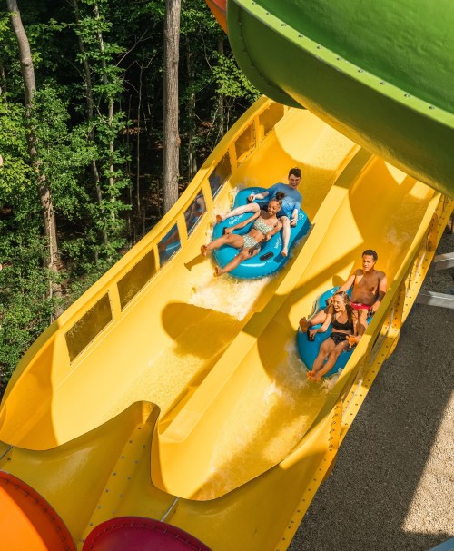 Two groups of people slide down a colorful water slide surrounded by trees on a sunny day.