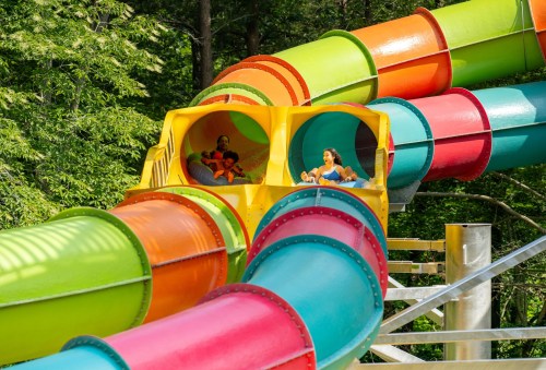 Two people slide down a colorful water slide surrounded by greenery, enjoying a fun day at a water park.