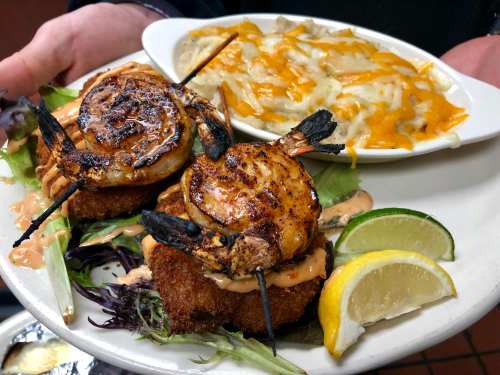 A plate featuring two grilled shrimp on breaded patties, served with creamy pasta and garnished with lime and lemon wedges.