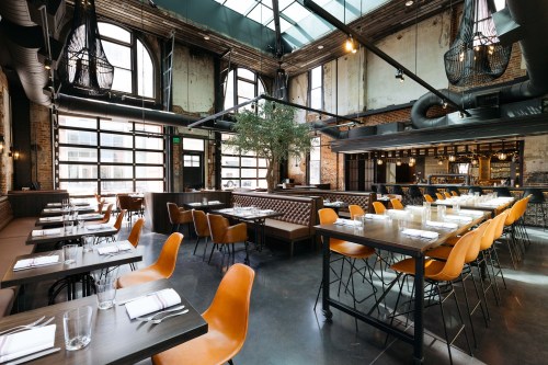 Modern restaurant interior with large windows, wooden tables, orange chairs, and industrial decor. Bright and spacious atmosphere.