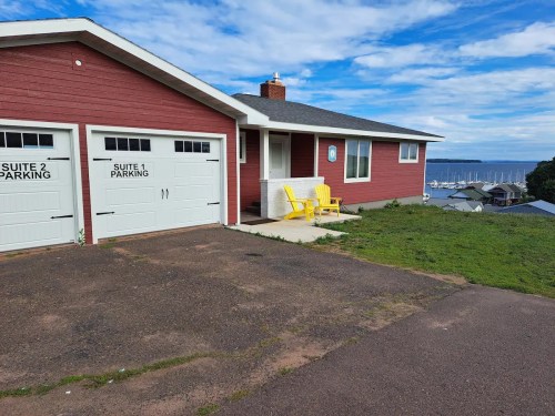 A red house with two garage doors, yellow chairs on the porch, and a view of the water and boats in the background.