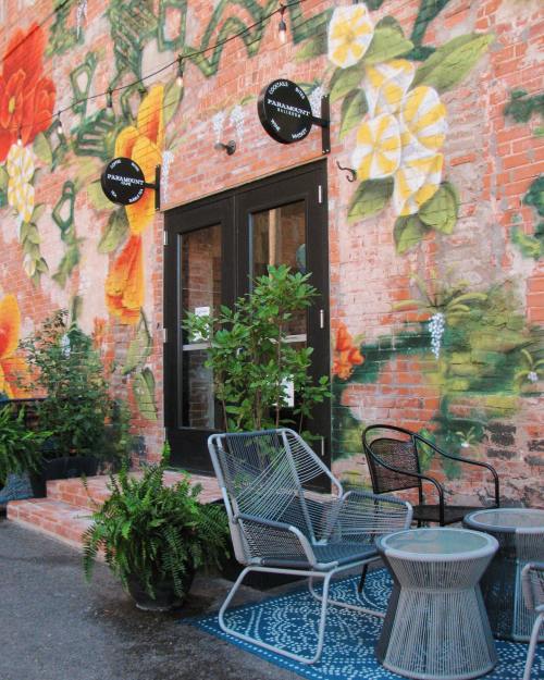A cozy outdoor seating area with plants, a patterned rug, and a colorful mural on a brick wall.