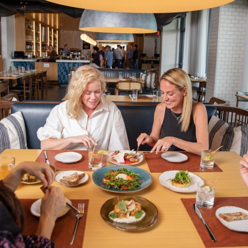 Two women enjoy a meal at a restaurant, sharing dishes and smiling at each other, with a variety of food on the table.