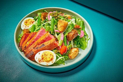 A vibrant salad with greens, cherry tomatoes, boiled eggs, crispy potatoes, and slices of grilled meat in a bowl.