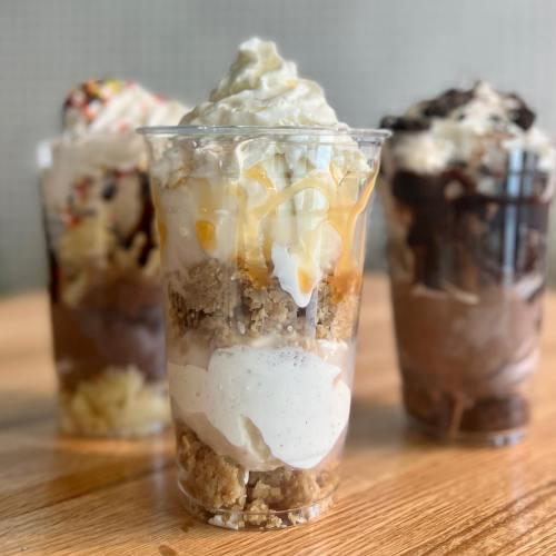 Three layered dessert cups with whipped cream, caramel, chocolate, and cookie crumbles on a wooden table.