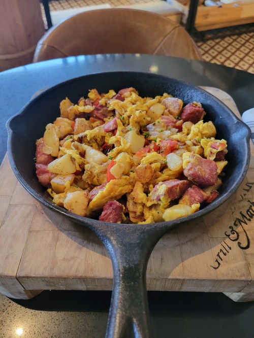 A cast iron skillet filled with scrambled eggs, diced potatoes, and chunks of meat, served on a wooden board.