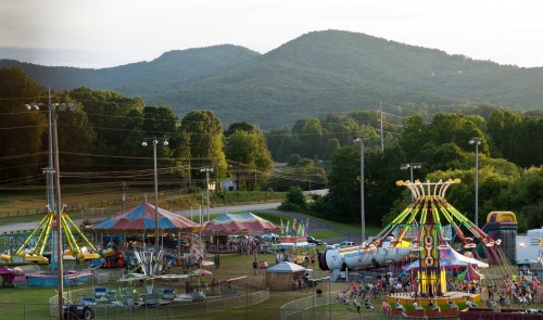 A vibrant carnival scene with rides and tents, set against a backdrop of rolling green mountains.