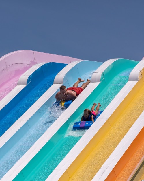 A man and a child slide down colorful water slides on a sunny day, splashing into the water below.
