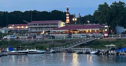 A lakeside scene featuring a red and white lighthouse, a motel, and boats docked at sunset.