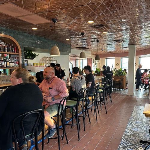 A cozy restaurant interior with patrons seated at a bar, plants, and a stylish ceiling. Natural light from windows.