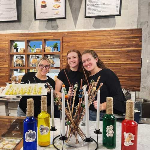 Three smiling women stand behind a counter with colorful bottles and decorative sticks, showcasing a variety of treats.