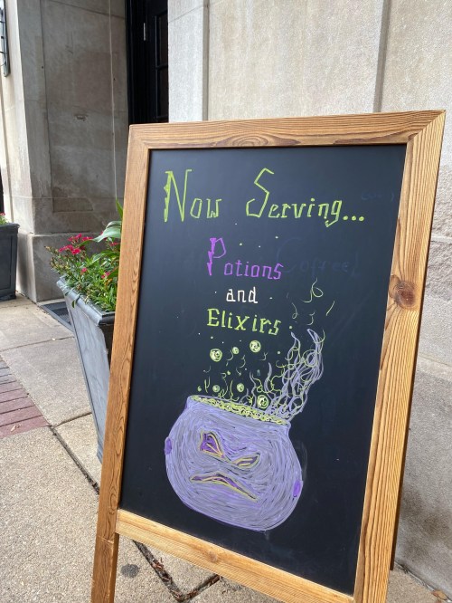 A chalkboard sign outside a building reads "Now Serving... Potions and Elixirs" with a drawing of a cauldron.