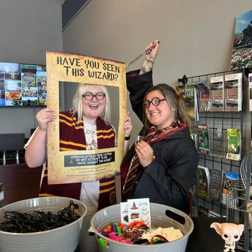 Two women in wizard costumes hold a "Have you seen this wizard?" sign, surrounded by craft supplies and decorations.