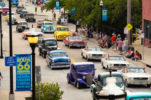 A vibrant parade of classic cars drives through a street lined with spectators and Route 66 festival banners.