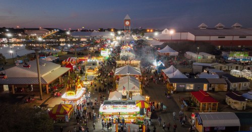Aerial view of a bustling fairground at dusk, with colorful stalls, lights, and crowds enjoying the festivities.