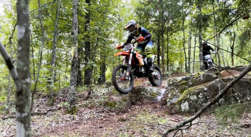A motocross rider navigates rocky terrain in a forest, with another rider visible in the background.