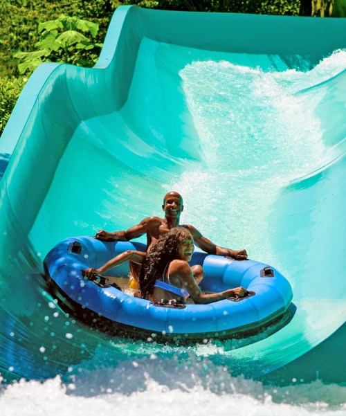 A man and woman enjoy a water slide, sitting on a blue inner tube as they splash down a vibrant green slide.