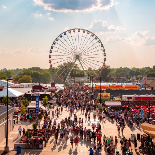 A bustling fairground scene with a large Ferris wheel, food stalls, and crowds enjoying the festivities under a sunny sky.