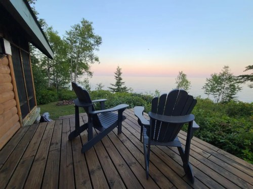 Two wooden adirondack chairs on a deck overlooking a serene lake at sunset, surrounded by trees.