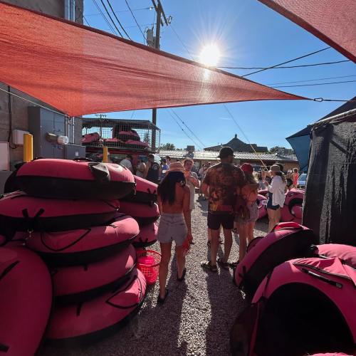 A sunny outdoor scene with people gathered near pink inflatable tubes and colorful tents, surrounded by power lines.