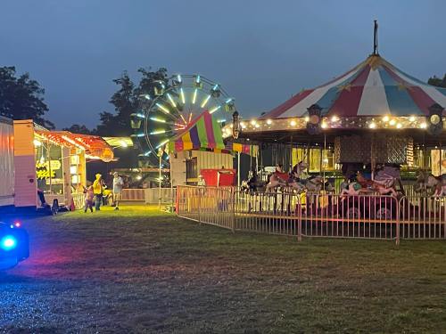 A carnival scene at dusk featuring a Ferris wheel, carousel, and colorful tents with lights.
