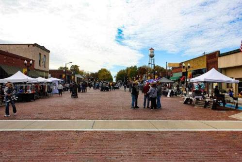A bustling street market with vendors under tents, people browsing, and a cloudy sky overhead.