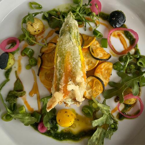 A beautifully plated dish featuring a stuffed zucchini blossom, surrounded by colorful vegetables and drizzled sauces.