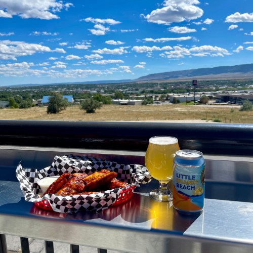 A plate of wings and a beer sit on a table with a scenic view of hills and a blue sky filled with clouds.