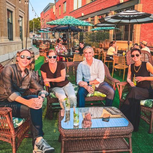 A group of four people relaxes outdoors at a café, enjoying drinks on a sunny day with others in the background.