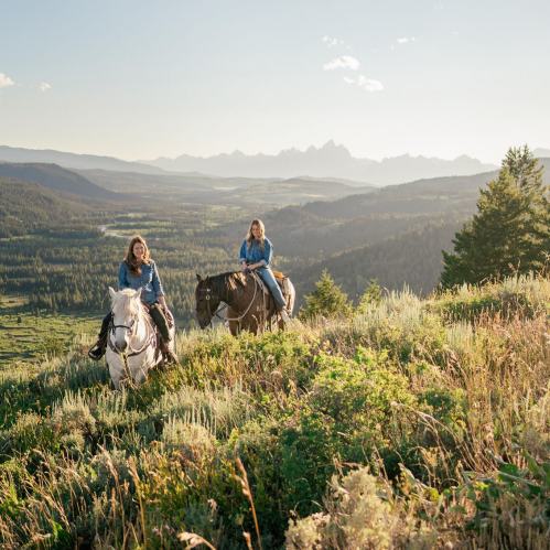 Two women on horseback ride through a scenic landscape with mountains and valleys under a clear sky.