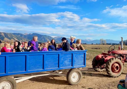 A group of children and adults ride in a blue wagon pulled by a red tractor, set against a scenic mountain backdrop.
