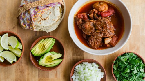 A bowl of meat stew with tomatoes, surrounded by tortillas, lime wedges, avocado slices, chopped onion, and cilantro.