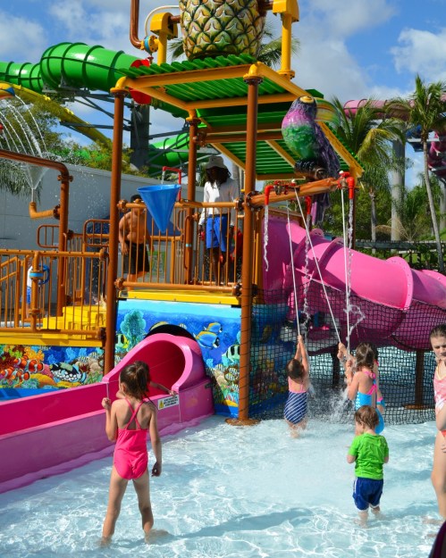 Children play in a water park area with slides, a splash pad, and colorful decorations under a sunny sky.